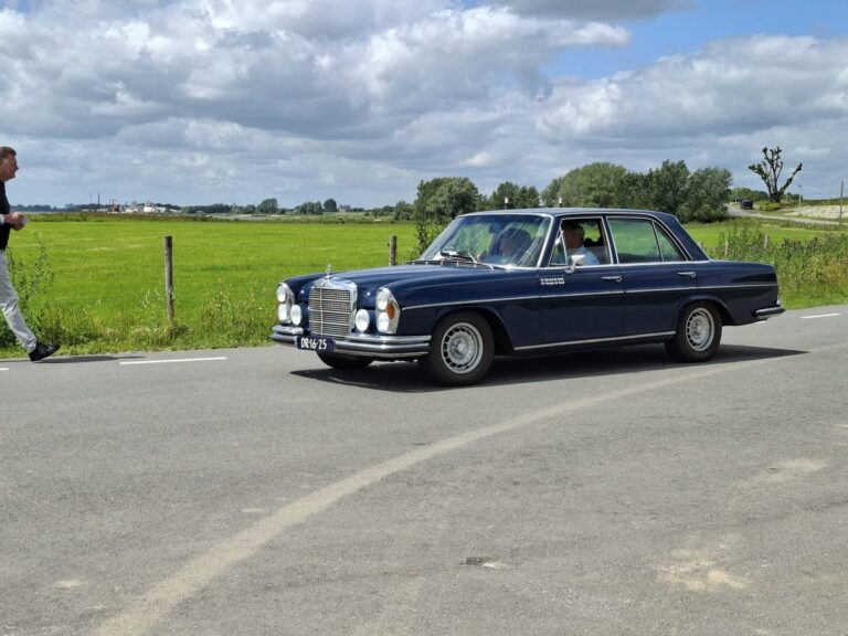 Een klassieke Mercedes rijdt over een dijkweggetje door de polder